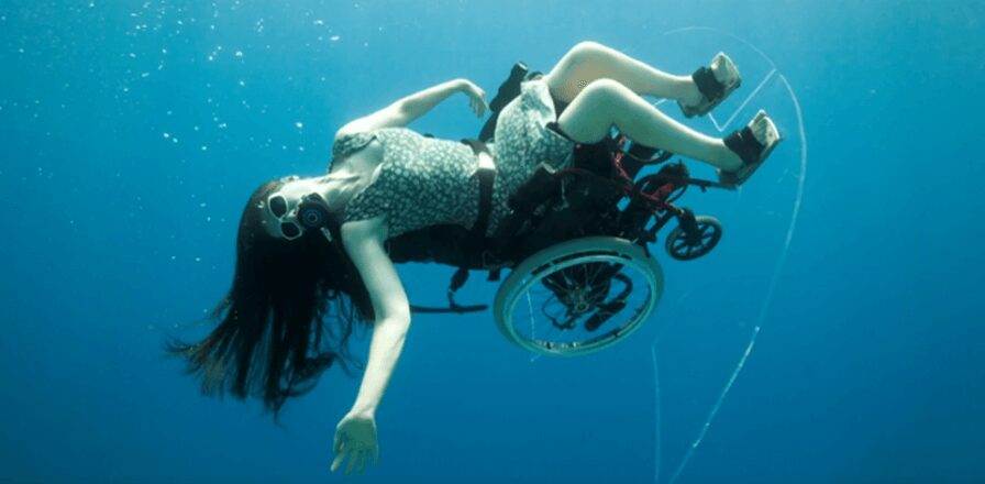 Sue Austin, Creating the Spectacle! &mdash; the artist floats underwater in a patterned dress and scuba mask, tilted almost horizontal in a wheelchair fitted with clear acrylic fins, her dark hair and one arm streaming out behind her.