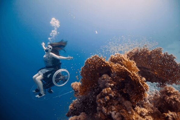 Sue Austin sits upright in her underwater wheelchair, one arm reaching forward as she passes a large orange coral head surrounded by a school of small fish, bubbles rising above her.
