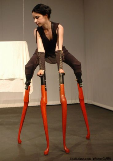Lisa Bufano, One Breath is an Ocean for a Wooden Heart &mdash; the artist perches on four tall orange stilts shaped like turned table legs, two on her arms and two on her legs, body folded forward and head lowered in a bare gray studio.