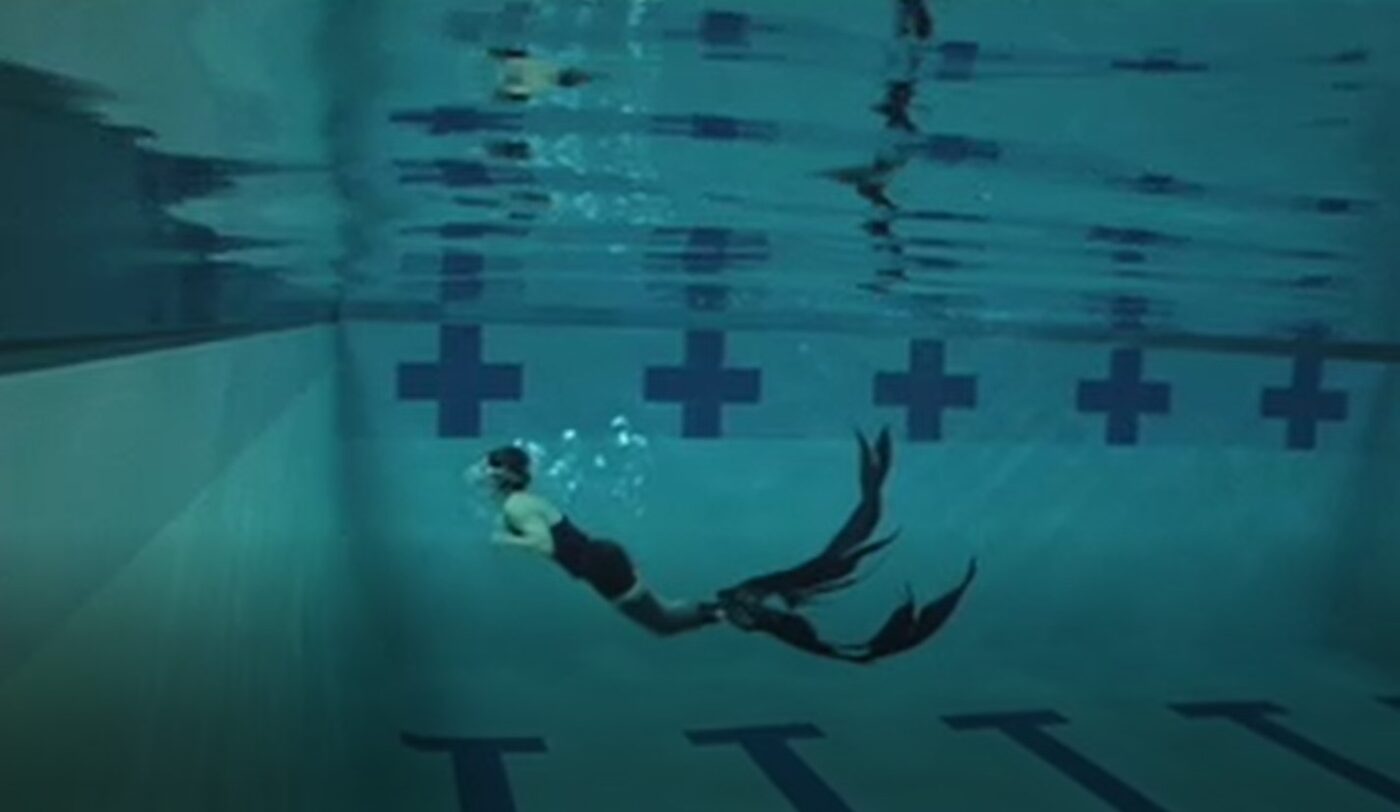 A performer glides horizontally through a swimming pool in goggles, trailing bubbles, a long dark feathery tail fin attached to her lower body.