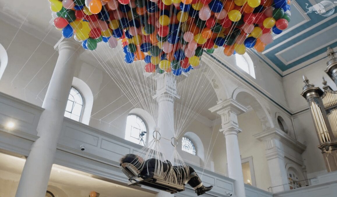 No&euml;mi Lakmaier, Cherophobia &mdash; the artist lies face up on a narrow board hanging from hundreds of strings beneath a dense cloud of multicolored balloons, floating high inside a white neoclassical church with an organ and pale blue ceiling.