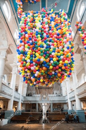 A vast mass of rainbow balloons fills the nave of a church, strings converging down to a small figure lying face up on a board, with visitors in the pews far below for scale.