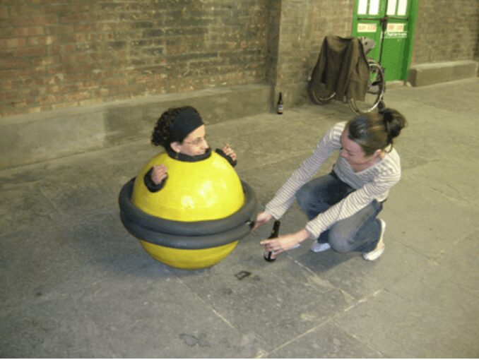 A performer is enclosed in a small yellow sphere ringed with black rubber; only her head and hands emerge. A second woman kneels beside her on the concrete floor of a brick-walled hall, reaching toward the sphere.