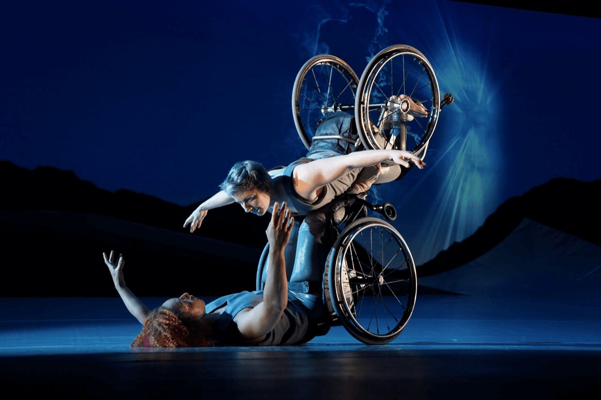 Alice Sheppard, DESCENT &mdash; Two dancers in a blue-lit duet: one lies on her back on the stage floor reaching upward, while the other balances her wheelchair tipped far back over her, leaning out with one arm extended and both wheels lifted. Dark hills and a burst of light fill the backdrop.