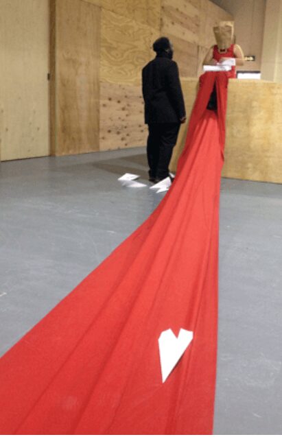 A seated performer with a paper bag over her head wears a red dress whose train runs the entire length of a plywood-walled room toward the viewer. White paper airplanes lie scattered across the fabric and floor; a figure in black stands with their back to the camera, facing her.